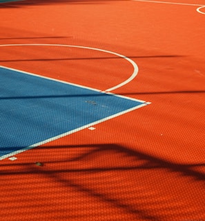 A vibrant sports court with sections of red and blue, featuring crisp white lines marking the boundaries. Shadows from a structure create geometric patterns across the surface, adding depth and interest to the scene.